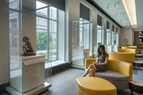 Woman reads a her phone while sitting on a yellow couch in front of a large window, nest to a statue of