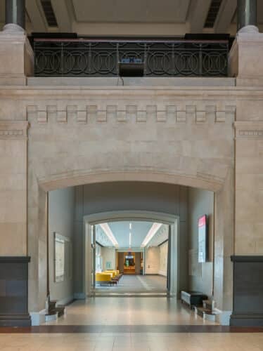 Entrance to the Emma Louise Schmidlapp wing of the Cincinnati Art Museum. Small groups of circular yellow couches line the right wall, each set in front of large windows letting in lots of light. There is a mural on the left wall., and an atrium entryway at the end of the corridor.