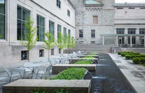 Courtyard at the Cincinnati Art Museum, featuring a row tables and chairs, Flanked by a row of trees on the left and small fountains that separate the table spaces on the right.