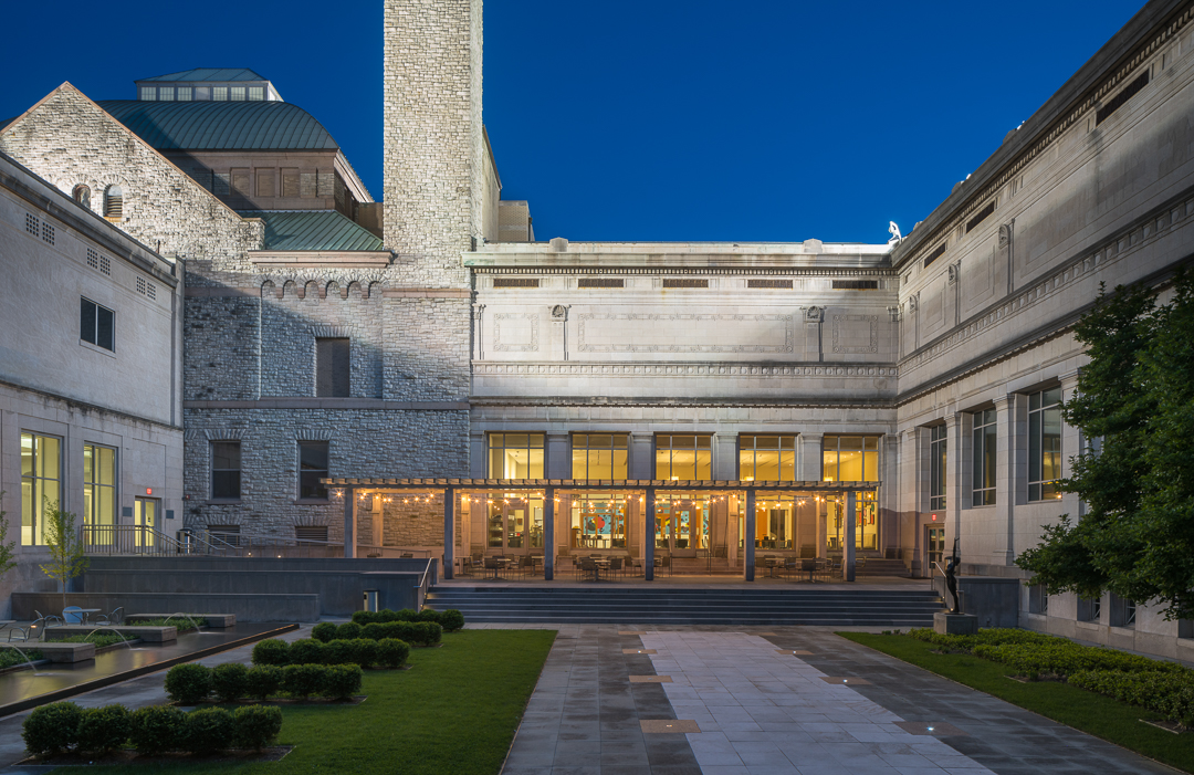 Night view of the courtyard at the Cincinnati Art Museum, featuring a row tables and chairs, flanked by a row of trees on the left and small fountains that separate the table spaces on the right.