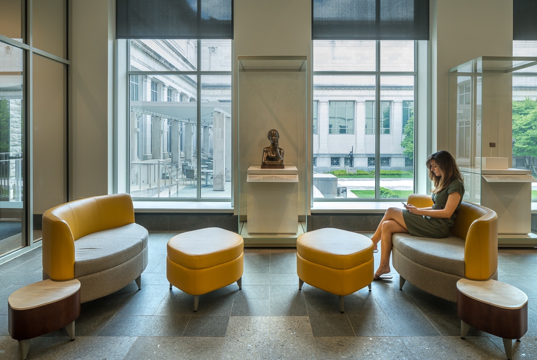 Woman reads a her phone while sitting on a yellow couch in front of a large window, next to a bust statue of Phillis Wheatley by Elizabeth Catlett.