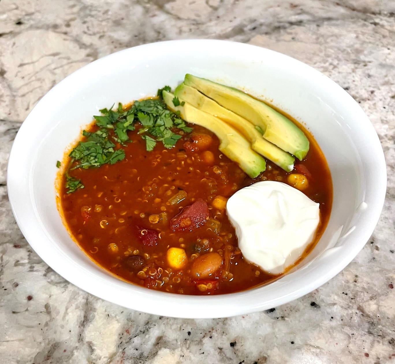 Photo of a bowl of Protien-Packed Veggie Chili with Quinoa, by Anna Milligan. She won the 2022 Best Looking Chili. Red with yellow corn throughout, topped with avocado, herbs, and sour cream.