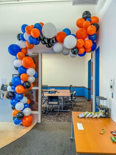 A balloon arch with orange, blue, black, and white balloons hangs over The Shelley conference room at SMBH's office.