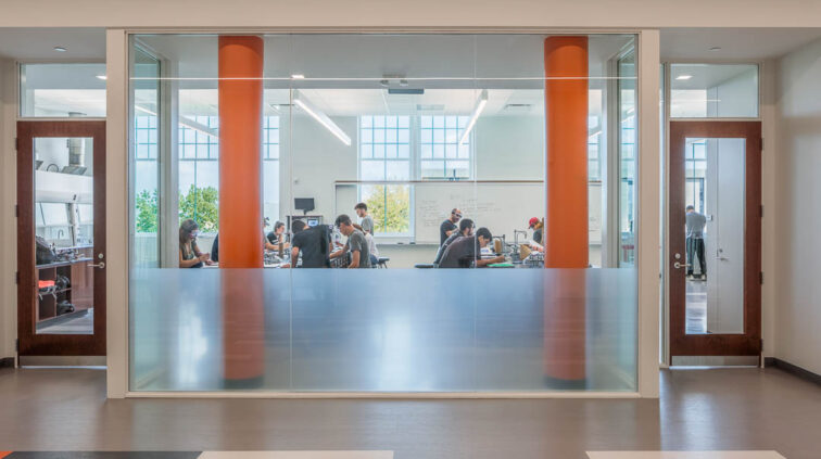 An interior view of a student work space inside Moseley Hall at Bowling Green State University. Room is shown behind a glass wall that looks between two large orange pillars, into the workspace.