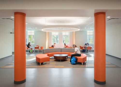An interior view of a student space inside Moseley Hall at Bowling Green State University. Room is shown between two large orange pillars, into the space, which includes a circular couth for large groups and smaller four-top tables for small student groups.