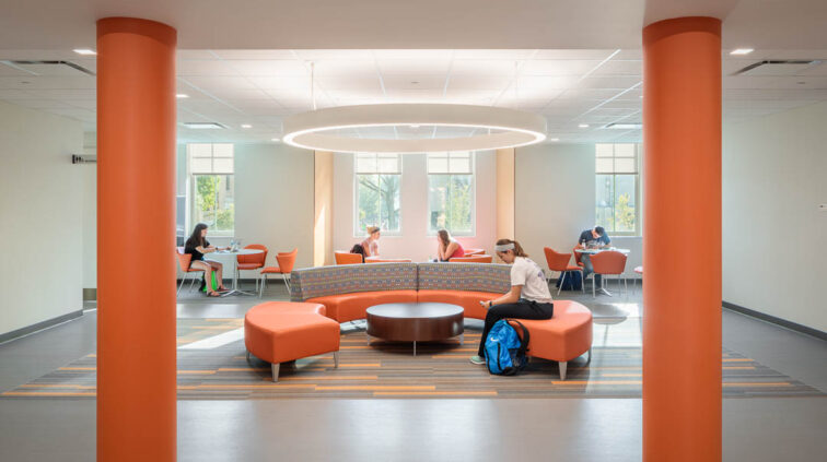 An interior view of a student space inside Moseley Hall at Bowling Green State University. Room is shown between two large orange pillars, into the space, which includes a circular couth for large groups and smaller four-top tables for small student groups.