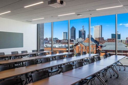 Interior photo of a classroom in Mitchell Hall at Columbus Community College. Photo features long tables that span the width of the classroom with a solid glass wall showcasing the downtown Columbus skyline.