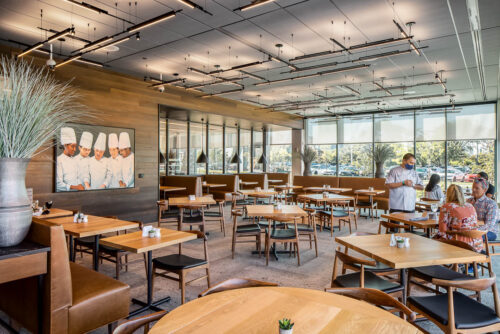 Interior photo of the student-run restaurant in the ground floor of Mitchell Hall at Columbus Community College. Features a room with floor-to-ceiling glass windows with natural wood toned tables and chairs arranged around the modern dining space.