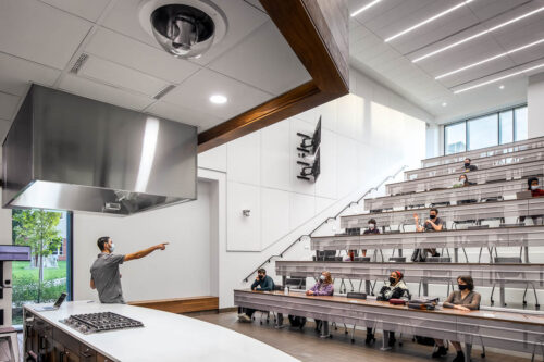 An interior view of the Teaching Kitchen Auditorium in Mitchell Hall at Columbus Community College. Features long desks for rows of students in an auditorium form so that all students can see. Features a kitchen with cameras and displays to show students what the instructor is doing up-close, even from the back of the room.