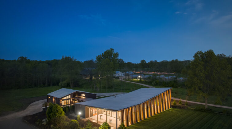 Exterior photo of The Gardens at Jorgensen Farms at dusk. The wood and glass structure is lit and glowing from within, against a blue sky in the background.