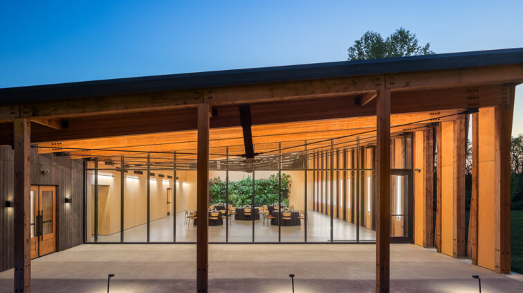 Exterior photo of The Gardens at Jorgensen Farms at dusk. The wood and glass structure is lit and glowing from within, against a blue sky in the background.