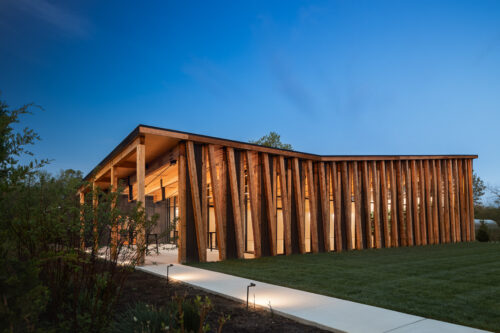 Exterior photo of The Gardens at Jorgensen Farms at dusk. The wood and glass structure is lit and glowing from within, against a blue sky in the background.
