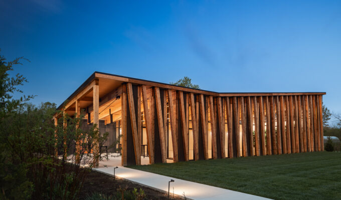 Exterior photo of The Gardens at Jorgensen Farms at dusk. The wood and glass structure is lit and glowing from within, against a blue sky in the background.