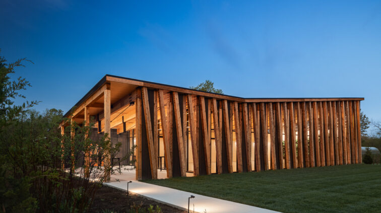 Exterior photo of The Gardens at Jorgensen Farms at dusk. The wood and glass structure is lit and glowing from within, against a blue sky in the background.