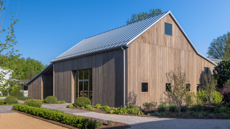 Exterior photo of The Gardens at Jorgensen Farms during the day. The wood and glass structure is lit with bright sunlight against a blue sky in the background.