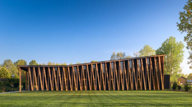 Exterior photo of The Gardens at Jorgensen Farms during the day. The wood and glass structure is lit with bright sunlight against a blue sky in the background.